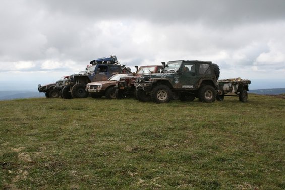 Several modified off-road vehicles and a trailer parked on a grassy hill under a cloudy sky