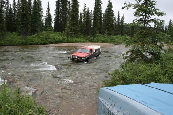 Red and grey pickup truck with camper shell crossing river; blue vehicle in foreground