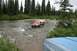 1990 Chevrolet Suburban - A red and grey pickup truck with a camper shell navigates a shallow river, with another vehicle partially visible in the foreground.