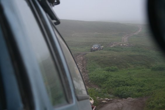 Two dark SUVs navigate a muddy track through a foggy, grassy landscape