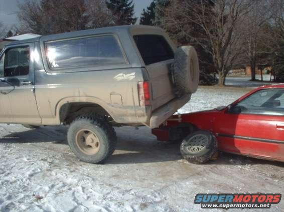 Ford Bronco is parked on the front end of a red car. Both vehicles are in a snowy area