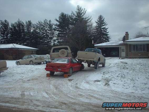 Red coupe on blocks with tan pickup truck behind it and tan SUV to the left
