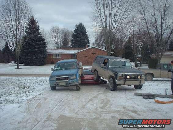 1986 Ford Bronco parked on a red car, with a blue GMC pickup and a tan pickup truck nearby in the snow