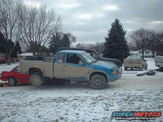 Blue and tan pickup truck parked next to a dark pickup truck, with a red car partially underneath the tan truck. Another tan car is in the background