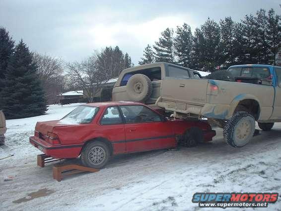 Red coupe on ramps, with a tan SUV on top and a blue pickup truck behind it in the snow