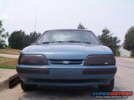 Front view of a blue 1987 Ford Mustang with tinted headlights and 'MUSTANG' text on the bumper