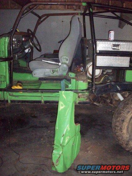 Modified off-road vehicle interior with roll cage, bucket seat, and detached green fender. Toolbox and beer can visible