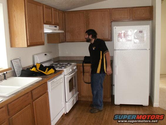 Man in kitchen looking at refrigerator. Wood cabinets, white stove, sink, and dishwasher visible