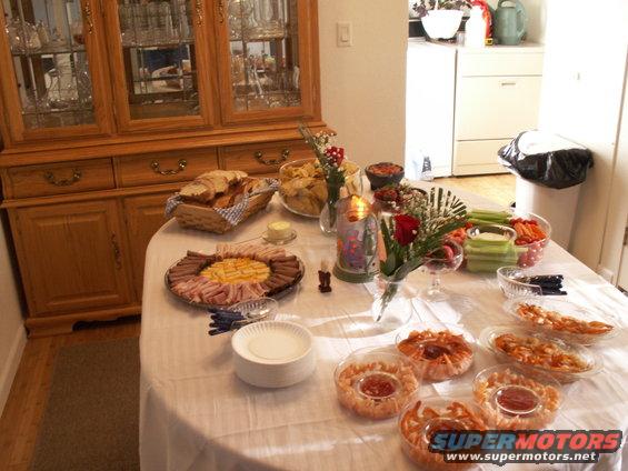 Buffet table with platters of meat, cheese, bread, chips, and vegetables, next to a wooden china cabinet
