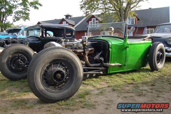 Green hot rod with exposed engine and wire wheels, parked next to a black car