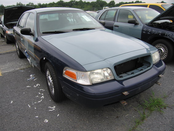 Front end of a dark blue and light blue 2001 Ford Crown Victoria with its grille missing. Another car with its hood up is to the left, and a police car is to the right