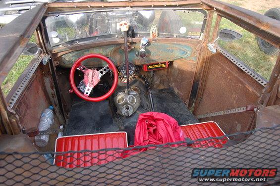 Interior of a rusted hot rod with red seats, red steering wheel, and a gas mask on the shifter