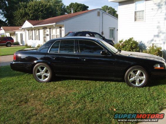 Black sedan with chrome wheels parked on grass, side view. Another vehicle is parked behind it