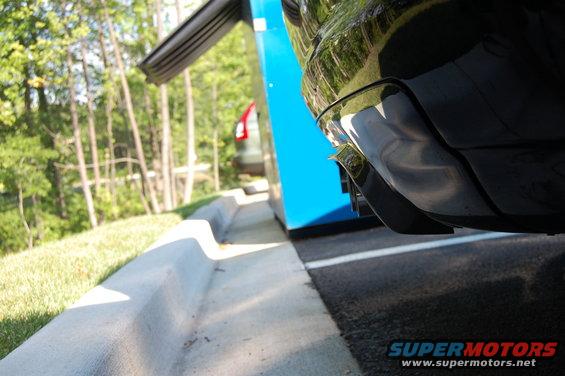 Close-up of a black car's rear bumper and exhaust pipes, with another car visible in the background