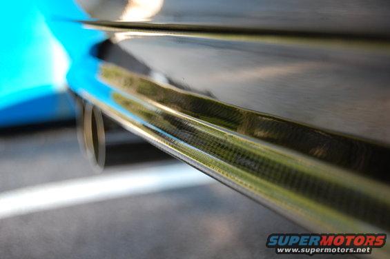 Close-up of a car's carbon fiber diffuser and exhaust pipe. A blue panel is visible on the left