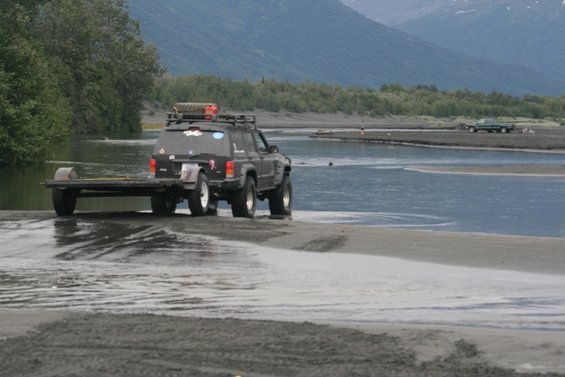 Black Jeep Cherokee XJ towing a trailer through shallow water, with another vehicle in the background