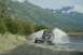 1990 Chevrolet Suburban - An SUV tows a flatbed trailer through a shallow body of water, kicking up spray against a backdrop of lush green mountains.