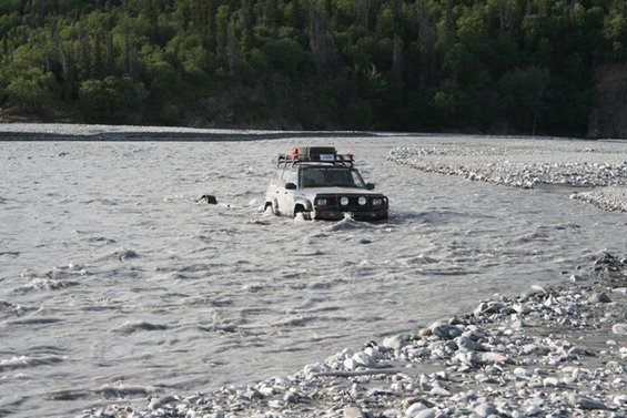 Off-road vehicle with roof rack driving through a shallow river with trees in background