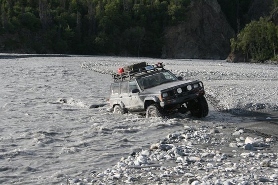 Jeep Cherokee XJ driving through a rocky riverbed with a roof rack loaded with gear