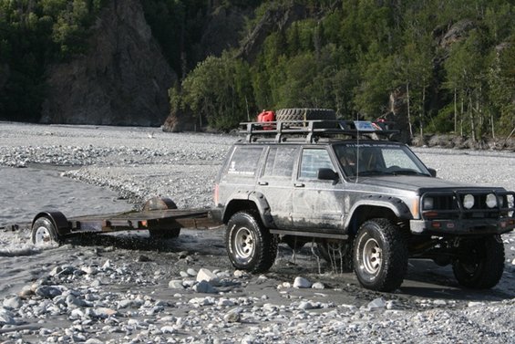 Gray Jeep Cherokee XJ towing a trailer through shallow water, with a roof rack carrying a spare tire and gas can
