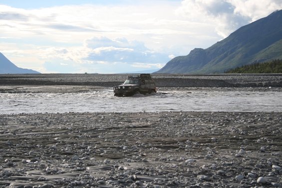 SUV with roof rack driving through a rocky riverbed with mountains in the background