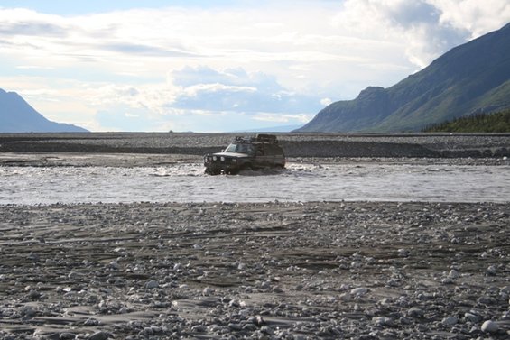 SUV with roof rack driving through shallow water in a rocky riverbed with mountains in the background