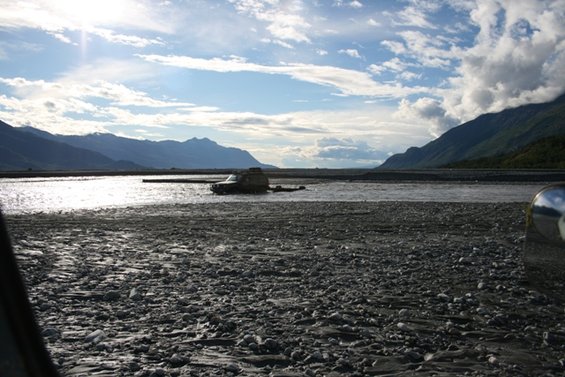Dark SUV partially submerged in a wide, shallow river with a rocky bed and mountains in the background