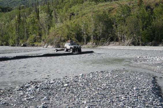 SUV towing a trailer through a shallow riverbed with trees in the background