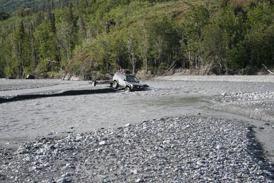4x4 vehicle with trailer stuck in a shallow river, trees and rocky banks in background