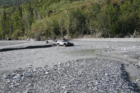 Gray SUV towing a trailer through a rocky riverbed with trees in the background
