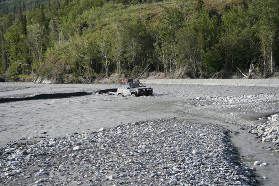 Gray SUV driving through a shallow riverbed with trees in the background