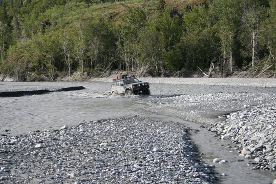 Off-road vehicle driving through a rocky riverbed with trees in the background. The vehicle appears to be a 1990s SUV