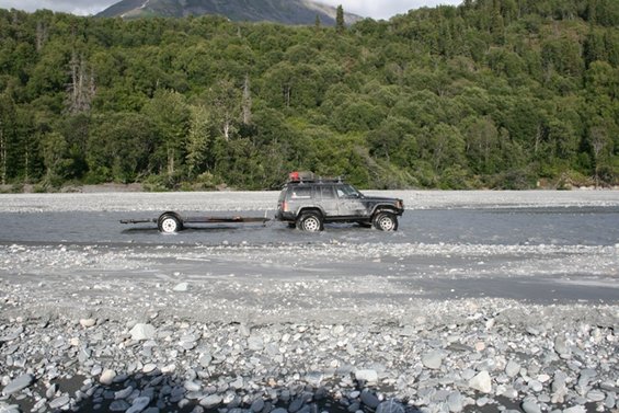 Jeep Cherokee with roof rack towing a small trailer through shallow river water, trees and mountain in background