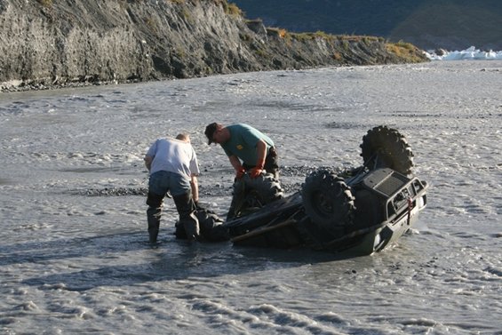 Two guys trying to right an ATV that's flipped over in a muddy riverbed