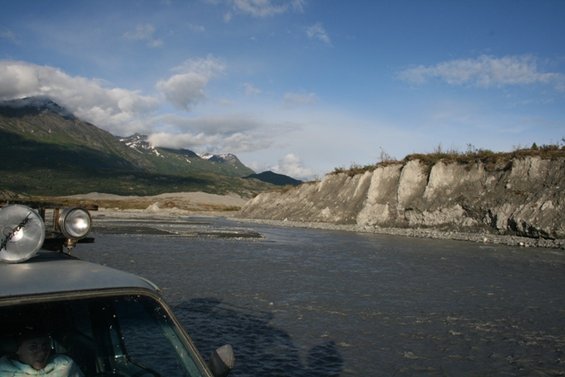 Front of an off-road vehicle with two large spotlights on the roof, parked in a wide, shallow riverbed with mountains in the background