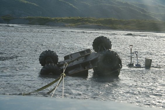 Off-road vehicle overturned in shallow river with two shovels and a bucket nearby