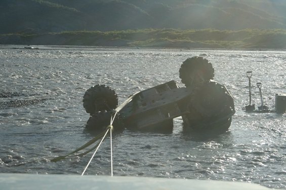 Tilted vehicle submerged in river, with large tires visible and a tow rope attached