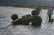 1990 Chevrolet Suburban - Two men attempt to recover an overturned ATV from a shallow riverbed, with a tow strap attached.