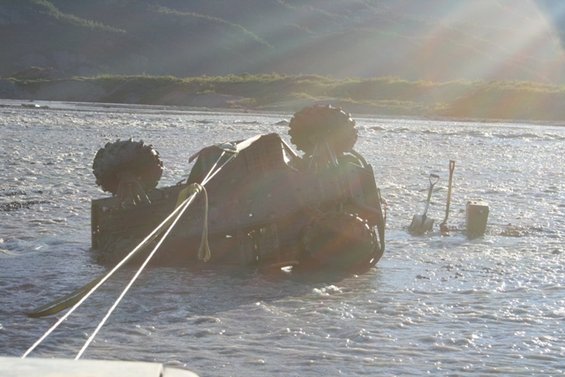 Overturned vehicle with large tires being pulled from shallow water by ropes; shovels and a bucket visible on shore