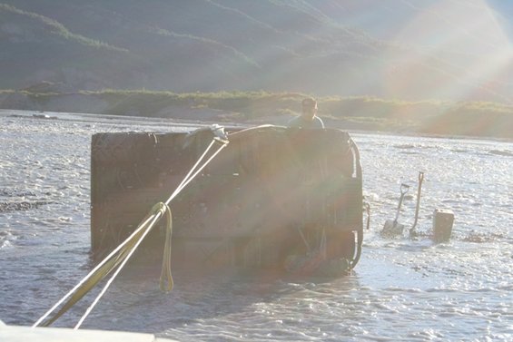 Vehicle partially submerged in a river, being pulled by ropes. Shovels are stuck in the riverbed nearby