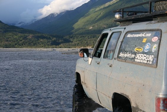 1990 Chevrolet Suburban parked in a shallow river with mountains in the background. Stickers on the rear window