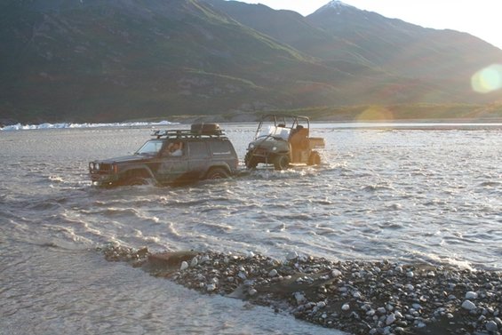 Dark SUV and utility vehicle crossing a shallow river with mountains in background