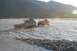 1990 Chevrolet Suburban - Two off-road vehicles navigate a wide, shallow riverbed under a bright, setting sun.