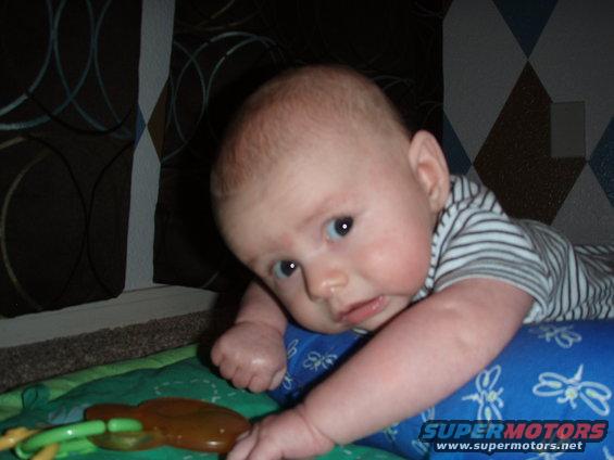 Baby lying on tummy on a playmat, looking at the camera. A toy is in front