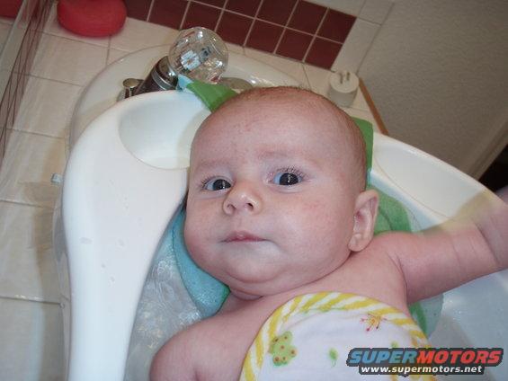 Baby in a white bathtub with a faucet and tiled wall. Watermark reads SUPERMOTORS