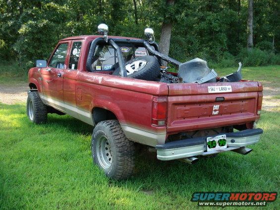 Red pickup truck with a roll bar and spare tire in the bed, parked on grass