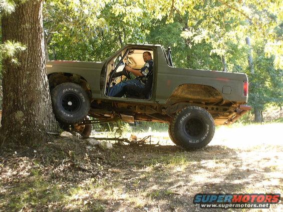 Olive green 1980 Ford Bronco with lifted suspension and off-road tires is partially on a tree trunk