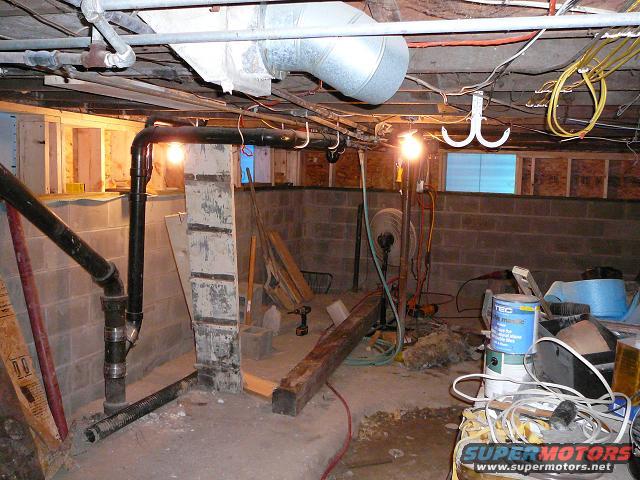 Basement with pipes, wires, concrete block walls, and construction debris. A fan and paint cans are visible