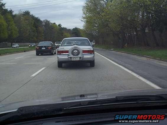 Rear view of a 1990s sedan with a spare tire on the trunk, driving on a highway. Another car is visible ahead