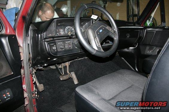 Interior of a red vehicle, showing dashboard, steering wheel, pedals, and seats. A man is visible in the background